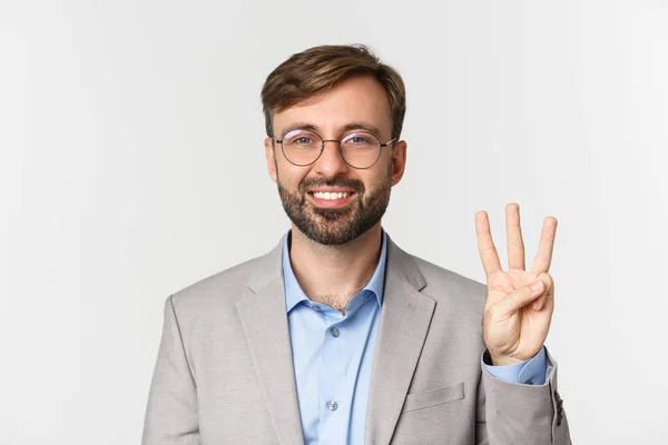 Close-up de homem barbudo bonito em óculos e terno cinza, mostrando o número três e sorrindo, de pé sobre fundo branco — Fotografia de Stock