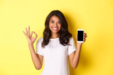Beyaz tişörtlü Afro-Amerikan güzel bir kızın portresi, tamam işareti ve akıllı telefon uygulaması gösteriyor, sarı arka planda duruyor.