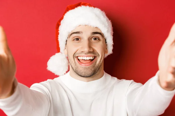 Gros plan d'un bel homme au chapeau de Père Noël, prenant un selfie ou ayant un appel vidéo, tenant la caméra avec les deux mains, debout sur un fond rouge — Photo