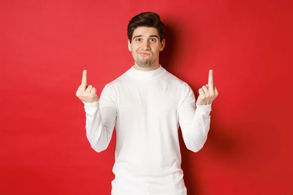 Image of pissed-off and distressed man telling to fuck off, showing middle-fingers and looking upset, standing over red background in white sweater — Stockfoto