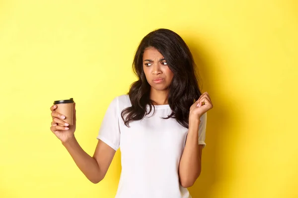 Portret van Afrikaans-Amerikaans meisje dat teleurgesteld kijkt naar beker, houdt niet van slechte koffie, staande over gele achtergrond — Stockfoto