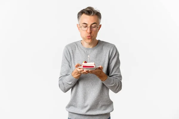 Retrato de homem de meia-idade atraente em camisola cinza e óculos, comemorando seu aniversário, soprando uma vela no bolo do dia b e fazendo o desejo, de pé sobre fundo branco — Fotografia de Stock