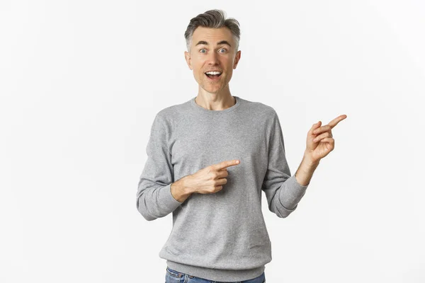 Bild av förvånad vacker medelålders man, leende och pekar finger rätt på logotyp eller promo banner, stående förvånad över vit bakgrund — Stockfoto