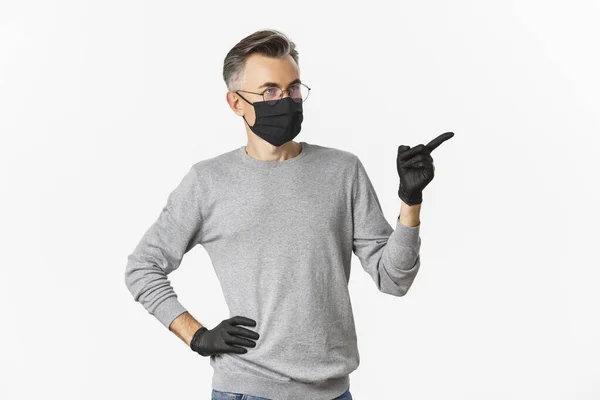 Concept de coronavirus, mode de vie et quarantaine. Homme d'âge moyen intrigué en masque médical, gants et lunettes pointant du doigt à droite et regardant la publicité, debout sur fond blanc — Photo