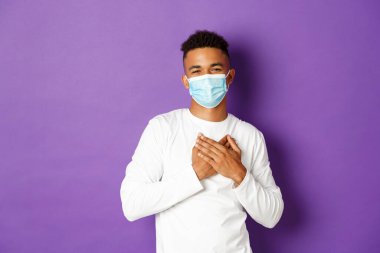 Koronavirüs, karantina ve yaşam tarzı kavramı. Tıbbi maske takmış yakışıklı Afro-Amerikan bir adamın resmi. Teşekkür ediyor, elleri yüreğinde, mor arka planda duruyor.