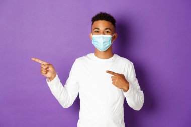 Koronavirüs, karantina ve yaşam tarzı kavramı. Tıbbi maskeli neşeli Afro-Amerikalı adam reklamını yapıyor, solu işaret ediyor ve gülümsüyor, mor arka planda duruyor.