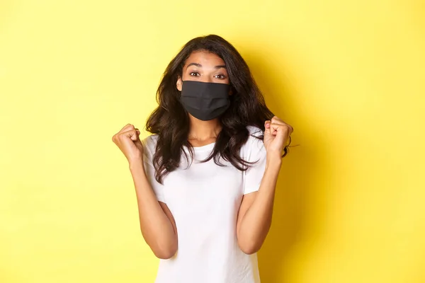 Concetto di covid-19, distanza sociale e stile di vita. Donna afro-americana allegra, indossando maschera nera, gioendo con le pompe pugno e sorridente, vincendo un premio, sfondo giallo — Foto Stock