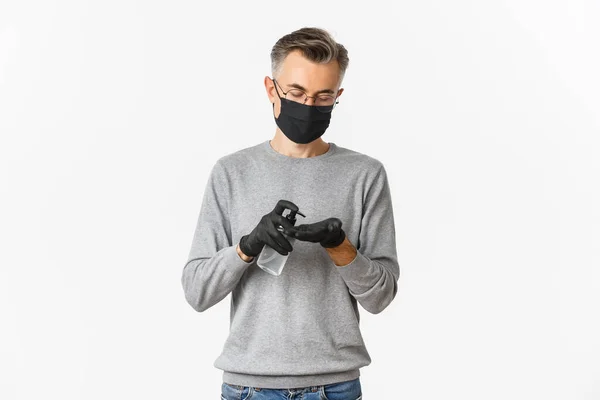 Concept of coronavirus, lifestyle and quarantine. Image of middle-aged man in medical mask and gloves, applying hand sanitizer, preventing catching covid-19, white background — Stock Photo, Image