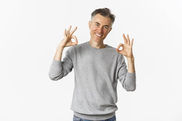 Portrait of successful handsome man in grey sweater, showing okay signs and smiling satisfied, approve something good, recommending product, standing over white background — Stock Photo, Image