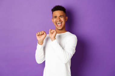 Beyaz kazaklı, zevkle dans eden, somurtan, başarıyı ve zaferi kutlayan mor arka planda mutlu bir şekilde duran aptal Afro-Amerikan erkek modeli.