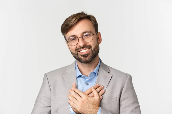 Close-up de empresário bonito em terno cinza e óculos, de mãos dadas no coração e agradecendo por algo, sorrindo grato, de pé sobre fundo branco — Fotografia de Stock