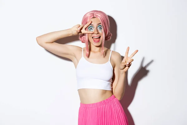 Spännande leende söt flicka i rosa peruk, visar fred tecken och ser förvånad, står i halloween kostym och rosa peruk — Stockfoto