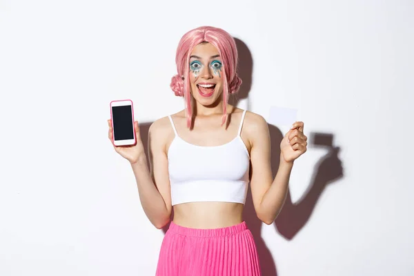 Wizerunek podekscytowanej dziewczyny w różowej peruce i stroju halloween, pokazujący kartę kredytową i ekran smartfona, uśmiechnięty szczęśliwy na białym tle — Zdjęcie stockowe