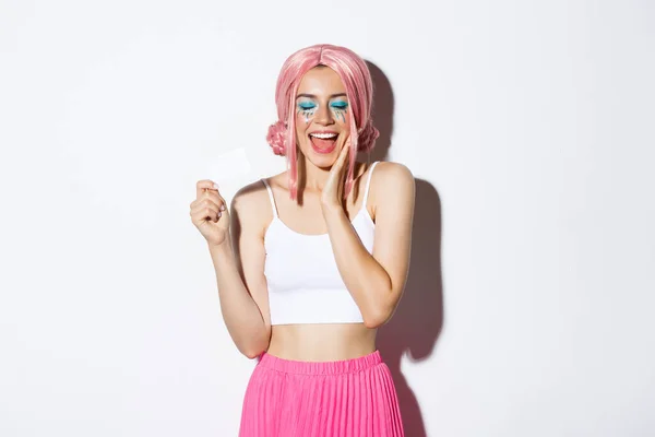 Porträt eines glücklichen, stylischen Mädchens mit rosa Perücke, geschlossenen Augen und Freudenschreien, das vom Einkaufen träumt und dabei seine Kreditkarte vorzeigt, das Outfit für die Halloween-Party trägt — Stockfoto