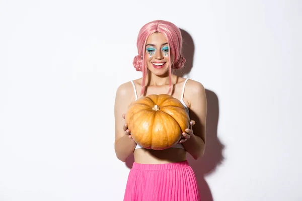 Porträtt av glad kvinna med rosa peruk och ljus makeup, ser glad på pumpa för halloween, står över vit bakgrund — Stockfoto