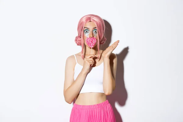 Bild av flirtig kvinnlig modell med rosa peruk och ljus makeup, kysser hjärtformade godis och ser förvånad, firar halloween — Stockfoto