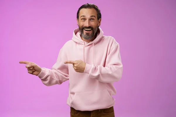 Charismatic happy good-looking mature 50s man wearing stylish hipster hoodie smiling excited pointing left index fingers impressed awesome interesting proposal standing pleased purple background — Stock Photo, Image