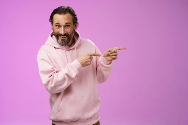 Sassy säker snygg macho man 50-talet i rosa hoodie leende brett inbjudande gå pekar höger visar intressant rolig plats umgås välkomnande ta en titt, stående lila bakgrund — Stockfoto