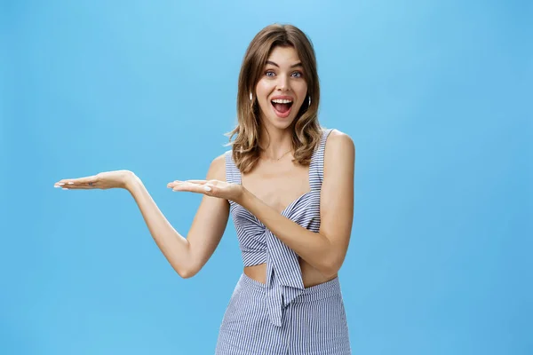 Woman presenting product over copy space. Good-looking charismatic adult tall woman with chestnut hair pointing at left side of wall and smiling broadly as if advertising against blue background