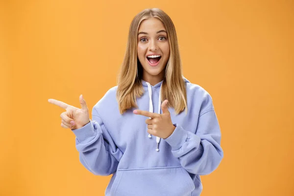 Amusant meisje wijzend naar nieuwsgierige kopieerruimte links met wijsvingers lachend opgewonden en verrast staand vermaakt met vrolijke grijns in knusse blauwe hoodie over oranje achtergrond — Stockfoto