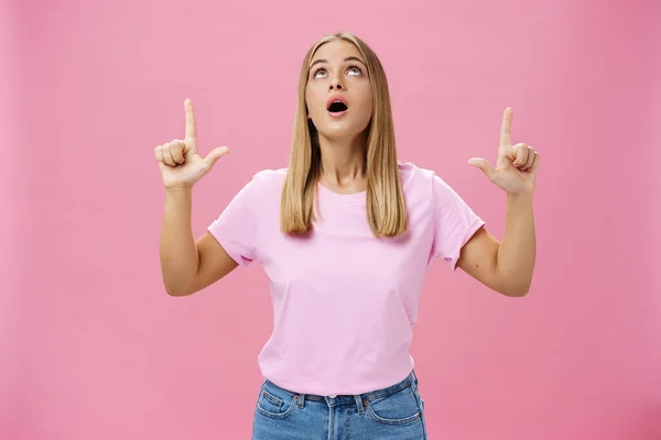 Retrato de mujer atractiva curiosa divertida e impresionada en camiseta dejando caer la mandíbula mirando y apuntando hacia arriba asombrado e intrigado mirando interesante objeto en el cielo posando sobre fondo rosa —  Fotos de Stock