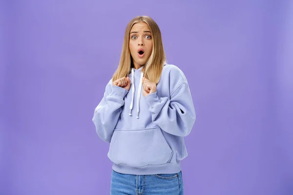 Woman scared of sudden loud noise in dark gasping looking shocked and concerned tilting backwards with frightened look pressing clenched fists to body from fear, wearing warm hoodie over purple wall — Stock Photo, Image