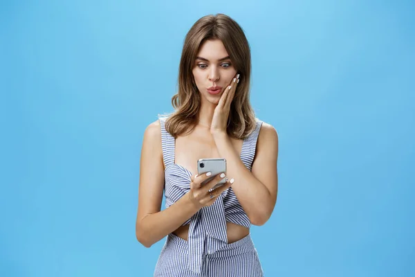 Женщина учится шокирующему слуху, получающему сообщение в социальной сети от друга, читающего новости со смартфона, складывающего губы, взволнованного, глядя на экран смартфона, прижимающего ладонь к щеке от изумления — стоковое фото