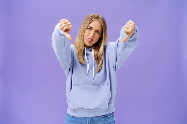 Indoor shot di moodie scontento e non impressionato fresco femminile moderna con i capelli biondi e la pelle abbronzata testa ribaltabile sporgente lingua mostrando pollici verso il basso in insoddisfazione posa contro muro viola — Foto Stock