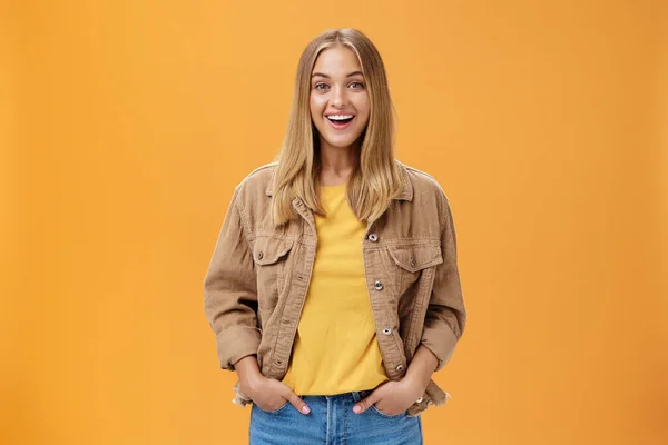 Charismatic tanned woman in corduroy jacket and yellow t-shirt ready for chilly autumn walk with friends smiling joyfully gazing entertained at camera holding hand in pockets casually over orange wall