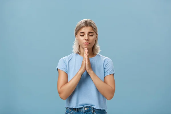 Chica europea amable y fiel de buen aspecto con el pelo rubio sonriendo lindo y tierno cogido de la mano en oración mientras pide deseo con suerte creyendo que Dios escucha sus oraciones con los ojos cerrados sobre la pared azul — Foto de Stock