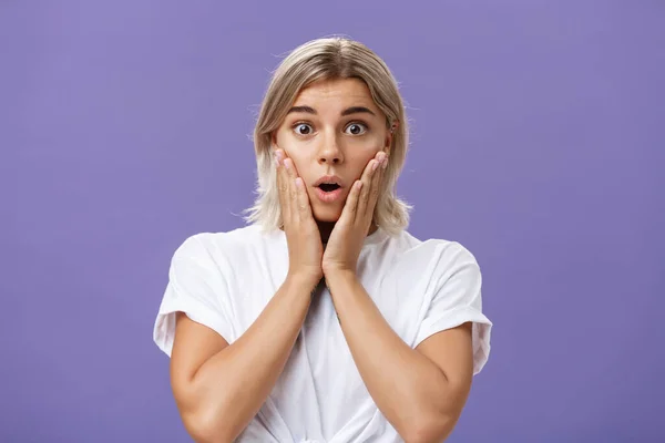 Waist-up shot of stunned impressed gorgeous tanned female in white t-shirt with blond hair gasping with opened mouth from surprise holding hands on cheeks from amazement and curiosity — Stock Photo, Image