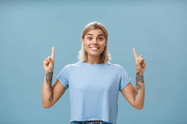 Tiro en la cintura de excitada y emocionada hermosa rubia feliz con peinado corto y tatuajes en los brazos sonriendo alegremente de asombro apuntando hacia arriba con los dedos índice levantados posando sobre fondo azul — Foto de Stock