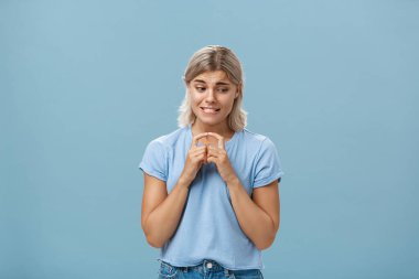 Kendine güveni olmayan, çekingen, sarı saçlı, dişlerini sıkan utangaç, kendinden emin olmayan ve mavi arka planda hoşlandığı çocuğa çıkma teklif eden aptal kız arkadaşının stüdyo fotoğrafı.
