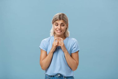 Studio shot of insecure and timid silly girlfriend with blond hair clenching teeth looking shy down and steepling index fingers unconfident trying ask out boy she likes over blue background clipart