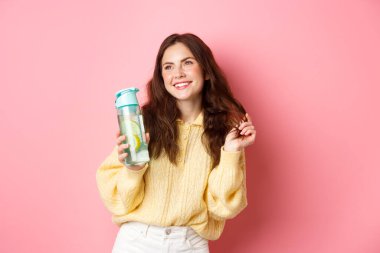 Güzel, çekici bir kadın saçlarıyla oynuyor ve gülümsüyor, yan bakıyor, su şişesinden sağlıklı bir içecek içiyor, pembe arka planda duruyor.