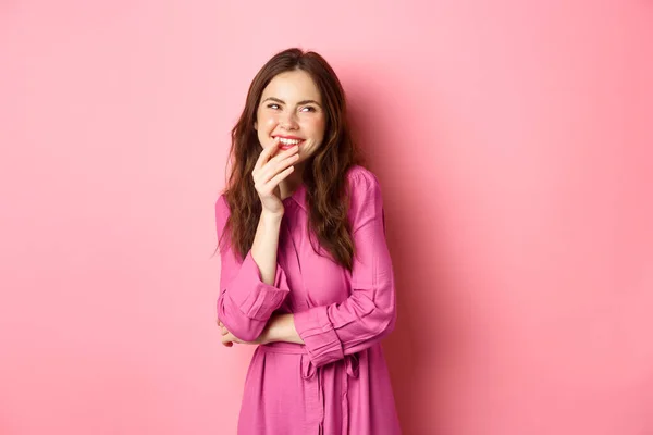 Coquettish young woman laughing and smiling, holding finger on lip wiht coy cute face, looking aside at promotional text, standing against pink background — Stock Photo, Image