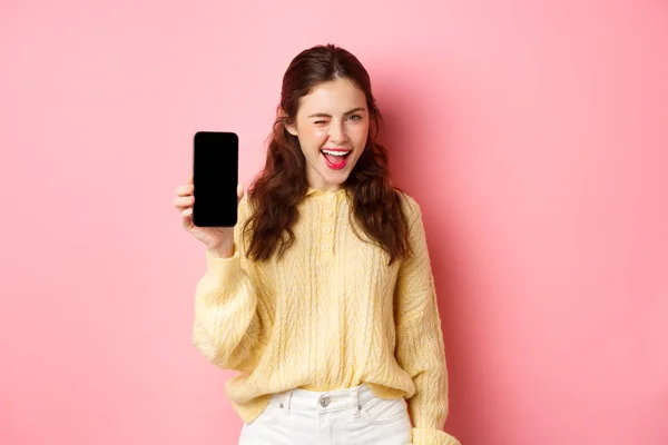 Изображение наглой привлекательной девушки, показывающей вам экран смартфона, подмигивающей и улыбающейся, рекомендующей мобильный телефон, стоящей на розовом фоне — стоковое фото