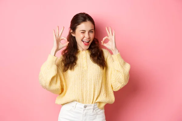 Retrato de menina bonita alegre piscando e dizendo sim, mostrando sinal ok, gesto certo, louvor bom trabalho, bem feito, de pé satisfeito contra fundo rosa — Fotografia de Stock