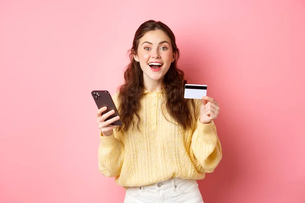 Tecnología y compras en línea. Chica emocionada haciendo orden, pagando en línea con tarjeta de crédito de plástico, sosteniendo el teléfono móvil y sonriendo a la cámara, fondo rosa — Foto de Stock