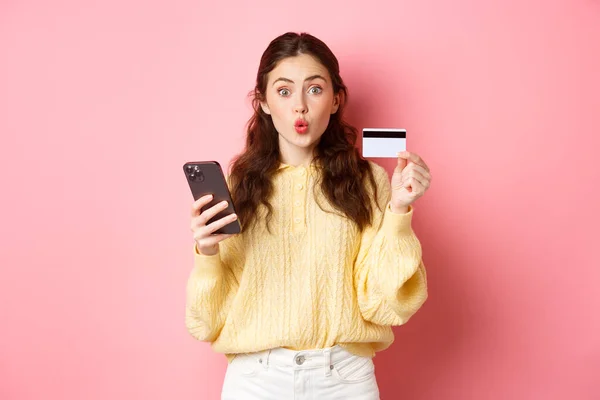 Tecnología y compras en línea. Emocionado comprador chica que muestra la tarjeta de crédito de plástico y la celebración de teléfono móvil, pagar con teléfono inteligente, orden en línea, de pie sobre fondo rosa — Foto de Stock