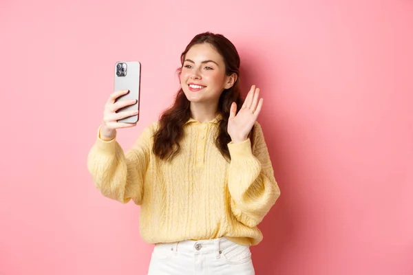 Porträtt av vänliga flicka talar på video chat app, viftande hand på smartphone kamera, har mobil konversation, står mot rosa bakgrund — Stockfoto