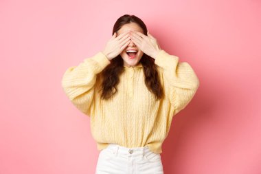 Heyecanlı genç doğum günü kızı gözlerini kapatıyor, şaşırmayı bekliyor, pembe arka planda gözleri bağlı duruyor.