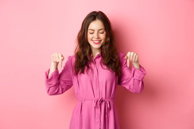 Bahar kıyafetleri içinde çekici bir kadın model, işaret ediyor ve aşağı bakıyor, telif alanını bir kenara koyuyor, pembe arka planda duruyor.