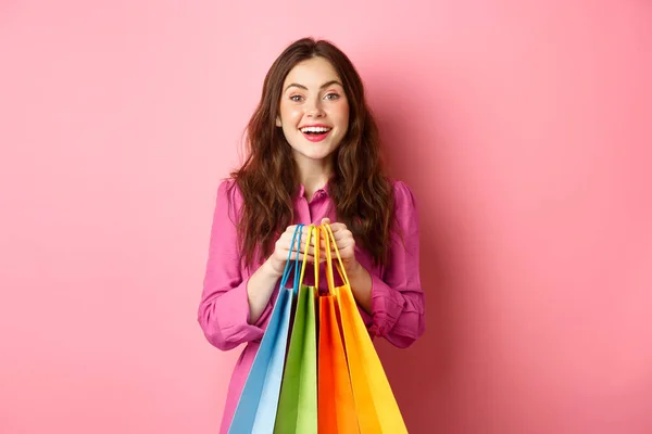 Junge schöne Frau geht einkaufen, hält Taschen in der Hand und lächelt aufgeregt, kauft mit Rabatten, steht vor rosa Hintergrund — Stockfoto