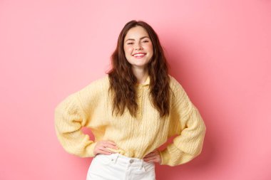 Güzel bir genç kadının portresi poz veriyor ve gülüyor, el ele tutuşuyor, sevinç ve pozitiflik hissediyor, cazibeli pembe arka planda duruyor.