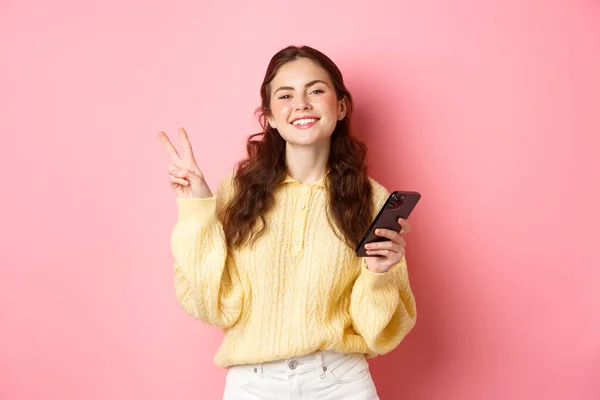 Tecnologia e compras online. Jovem feliz sorrindo satisfeito, segurando smartphone, mostrando v-sinal e olhar positivo para a câmera, de pé sobre fundo rosa — Fotografia de Stock