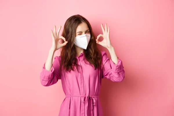 Covid-19, pandemic and lifestyle concept. Cheerful girl shows okay sign, wears medical respirator as preventive measure from corona, pink background — Stock Photo, Image