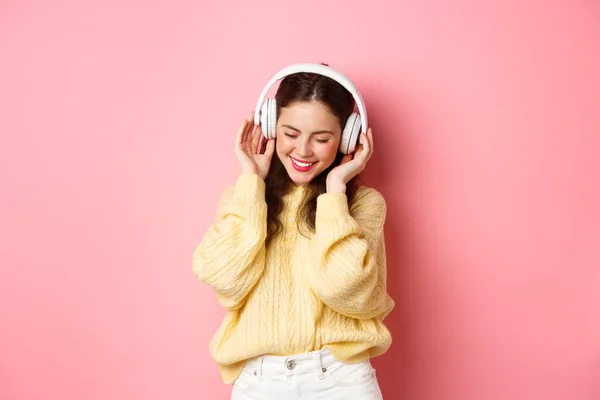 Retrato de chica morena atractiva disfrutando escuchando música, bailando y sonriendo complacido, de pie sobre fondo rosa — Foto de Stock
