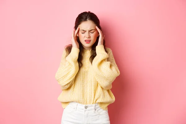 Mujer joven que tiene dolor de cabeza terrible, tocando la cabeza y la mueca por el dolor de migraña, necesita analgésicos, de pie contra el fondo rosa —  Fotos de Stock