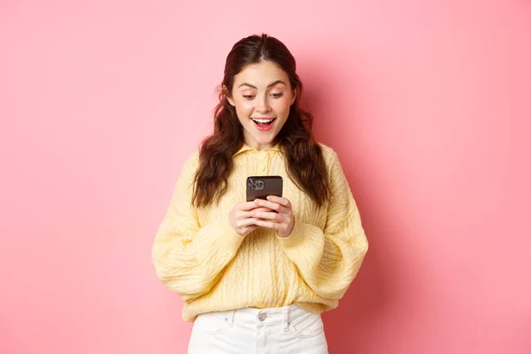 Портрет взволнованной брюнетки, задыхающейся от изумления, читающей сообщение на смартфоне, удивлённой на мобильный телефон, стоящей на розовом фоне — стоковое фото